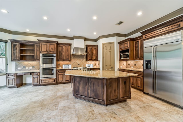 kitchen with light stone countertops, sink, wall chimney range hood, built in appliances, and an island with sink