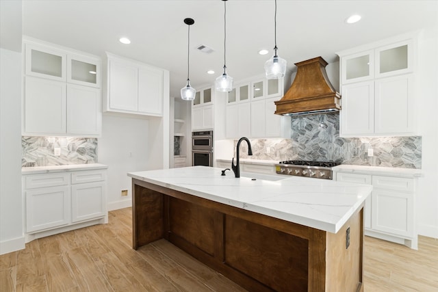 kitchen featuring decorative backsplash, premium range hood, a kitchen island with sink, sink, and white cabinetry