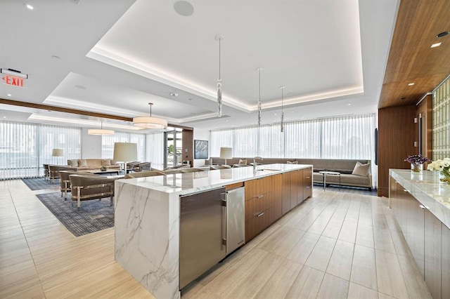 kitchen with light stone countertops, a large island with sink, a tray ceiling, and hanging light fixtures