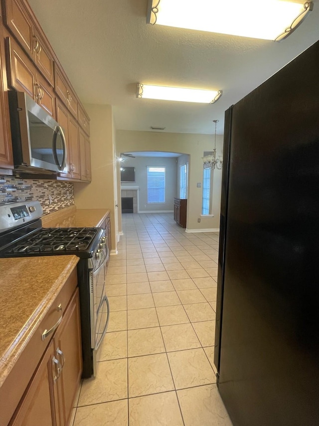 kitchen featuring a tile fireplace, stainless steel appliances, tasteful backsplash, light tile patterned flooring, and ceiling fan with notable chandelier