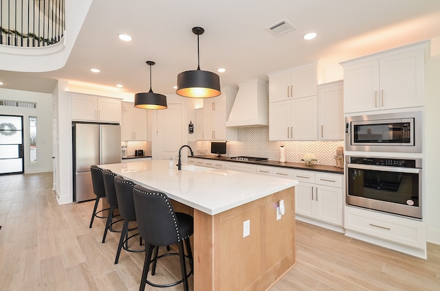 kitchen with white cabinets, premium range hood, appliances with stainless steel finishes, and an island with sink