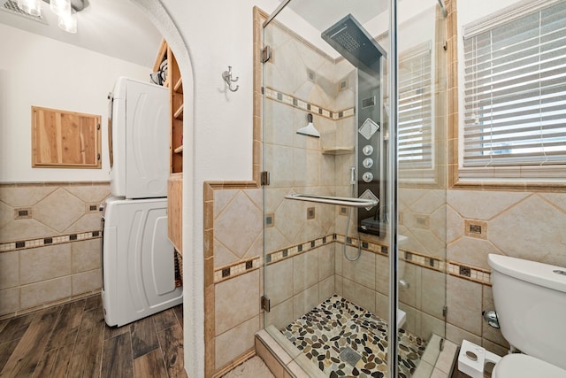 bathroom featuring stacked washing maching and dryer, a shower with door, wood-type flooring, tile walls, and toilet