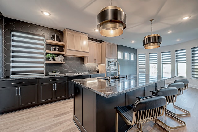 kitchen featuring sink, tasteful backsplash, decorative light fixtures, dark stone countertops, and a kitchen island with sink