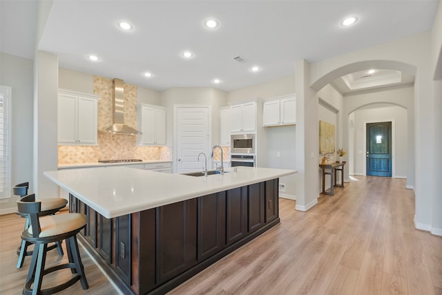 kitchen with light wood-type flooring, wall chimney exhaust hood, sink, a large island with sink, and white cabinetry