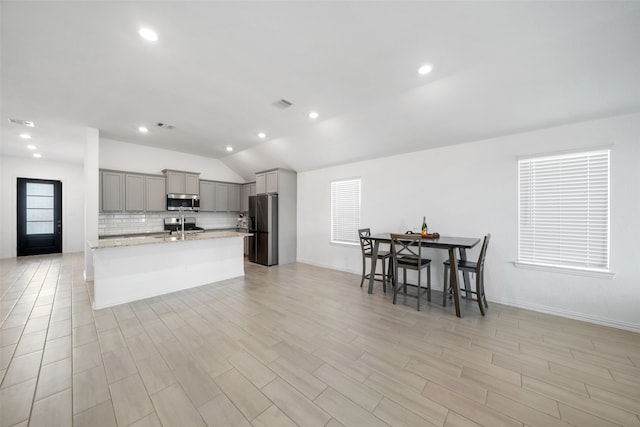 kitchen with a kitchen bar, gray cabinetry, stainless steel appliances, vaulted ceiling, and a center island with sink