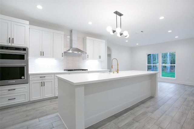 kitchen with decorative light fixtures, wall chimney range hood, white cabinetry, an island with sink, and stainless steel appliances