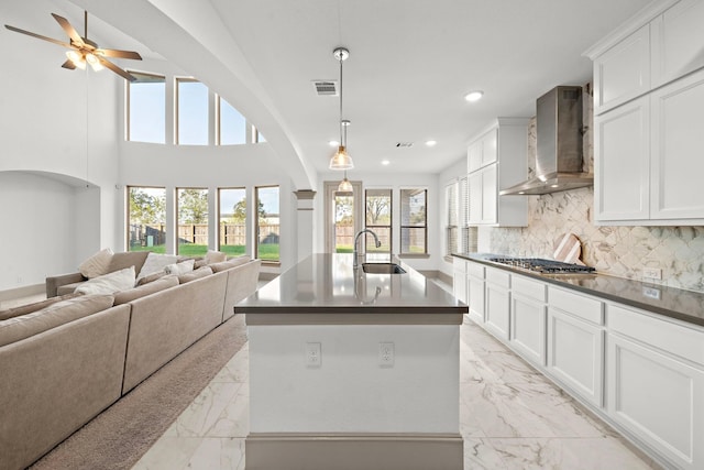 kitchen with a kitchen island with sink, stainless steel gas cooktop, sink, wall chimney range hood, and white cabinetry