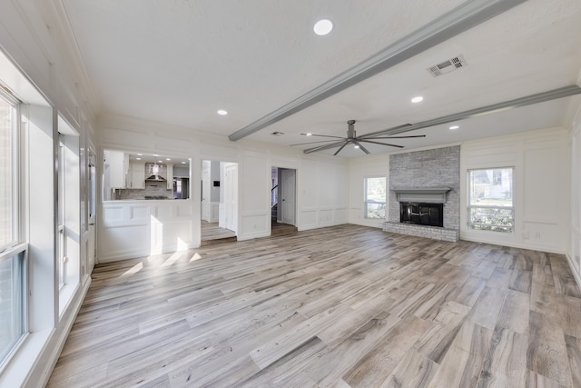 unfurnished living room featuring beam ceiling, ceiling fan, a brick fireplace, light hardwood / wood-style flooring, and ornamental molding