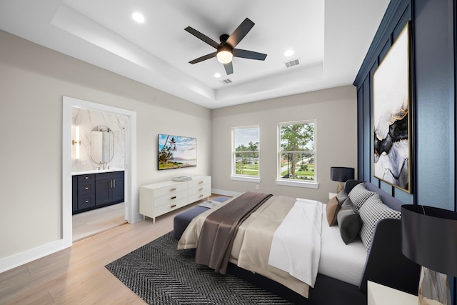 bedroom featuring ceiling fan, light hardwood / wood-style floors, a raised ceiling, and ensuite bath