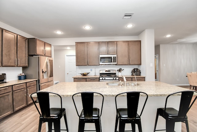 kitchen with light stone countertops, light wood-type flooring, stainless steel appliances, a kitchen island with sink, and sink