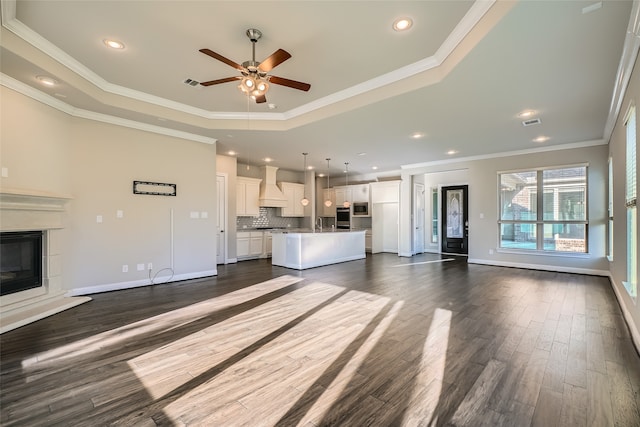 unfurnished living room with a tray ceiling, ceiling fan, dark wood-type flooring, and ornamental molding