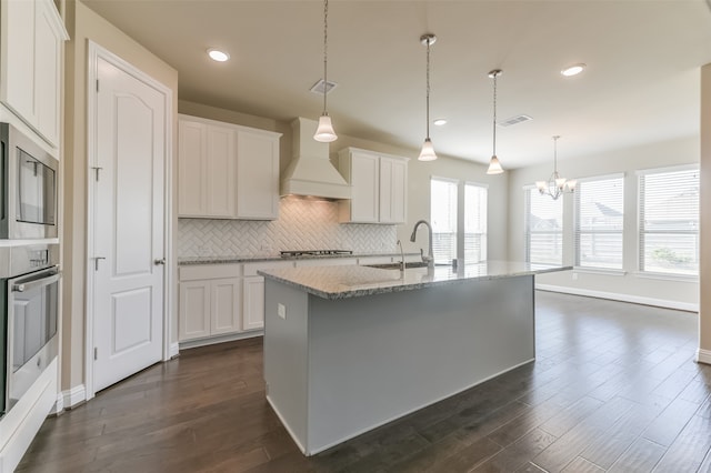 kitchen with premium range hood, stainless steel appliances, decorative light fixtures, white cabinets, and an island with sink