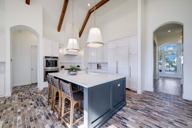 kitchen with high vaulted ceiling, an island with sink, decorative light fixtures, white cabinetry, and stainless steel double oven