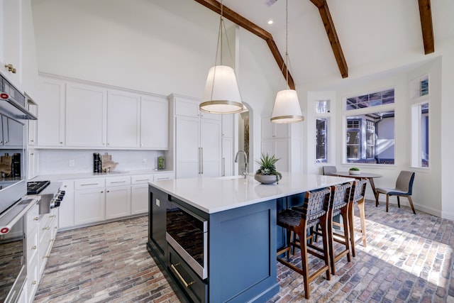 kitchen with beamed ceiling, an island with sink, decorative light fixtures, white cabinets, and appliances with stainless steel finishes