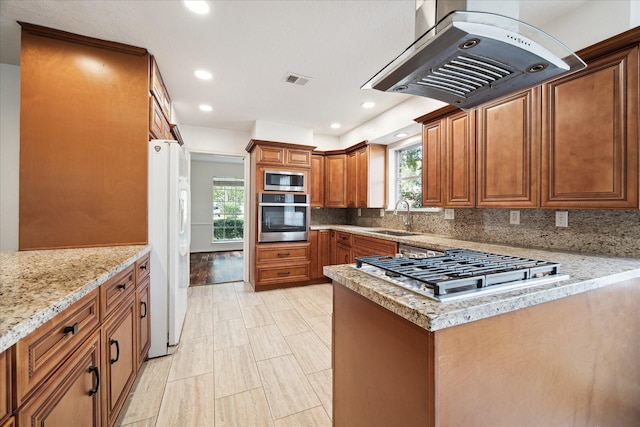 kitchen featuring tasteful backsplash, island range hood, appliances with stainless steel finishes, brown cabinets, and a sink