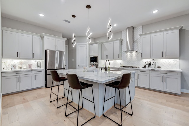 kitchen featuring wall chimney exhaust hood, pendant lighting, decorative backsplash, a center island with sink, and appliances with stainless steel finishes