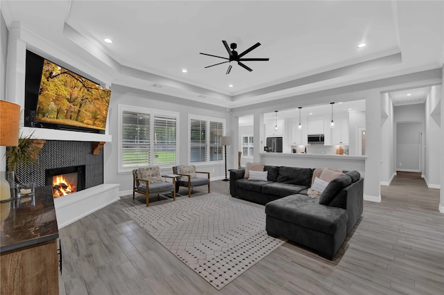 living room featuring light wood-type flooring, a raised ceiling, ceiling fan, and a tiled fireplace