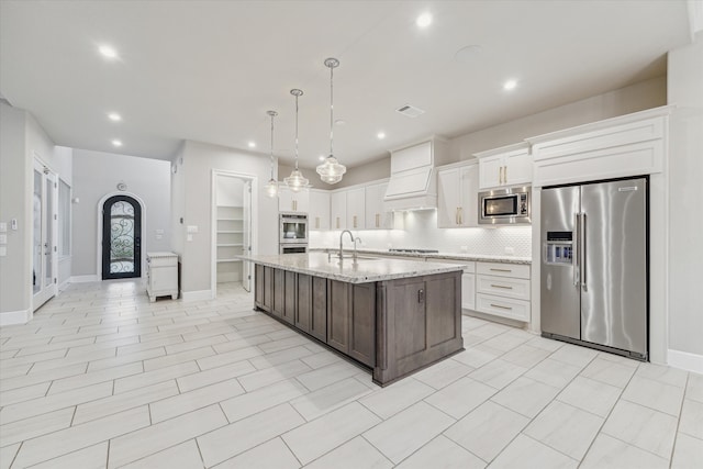 kitchen featuring white cabinets, hanging light fixtures, light stone countertops, an island with sink, and appliances with stainless steel finishes