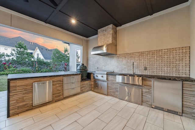 kitchen featuring stainless steel fridge, tasteful backsplash, dark stone counters, wall chimney range hood, and sink