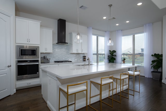 kitchen featuring white cabinets, wall chimney range hood, sink, an island with sink, and appliances with stainless steel finishes