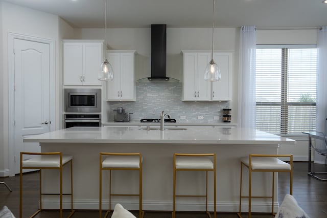 kitchen with light stone countertops, stainless steel appliances, a kitchen island with sink, wall chimney range hood, and white cabinets