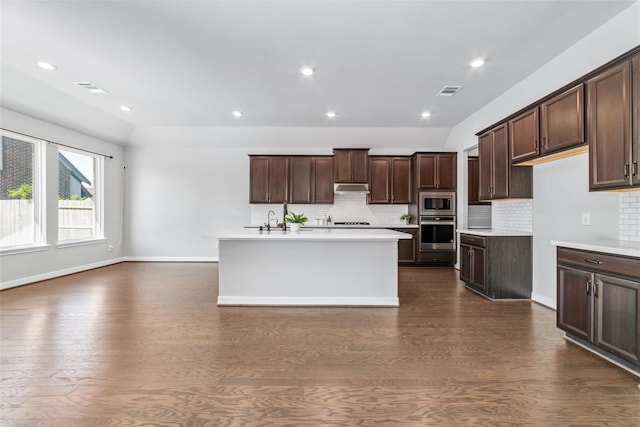 kitchen with dark wood-type flooring, an island with sink, appliances with stainless steel finishes, tasteful backsplash, and dark brown cabinets