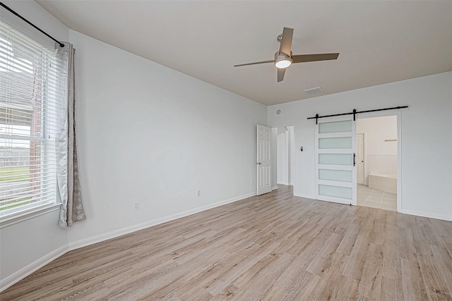 unfurnished bedroom with ceiling fan, a barn door, light wood-type flooring, and ensuite bath