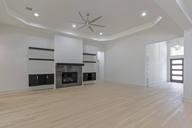 unfurnished living room with a raised ceiling, a tiled fireplace, light hardwood / wood-style flooring, and ceiling fan with notable chandelier