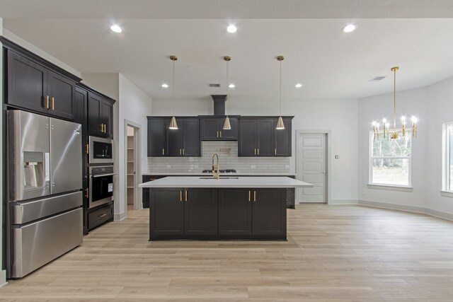 kitchen featuring appliances with stainless steel finishes, light wood-type flooring, a kitchen island with sink, pendant lighting, and a chandelier