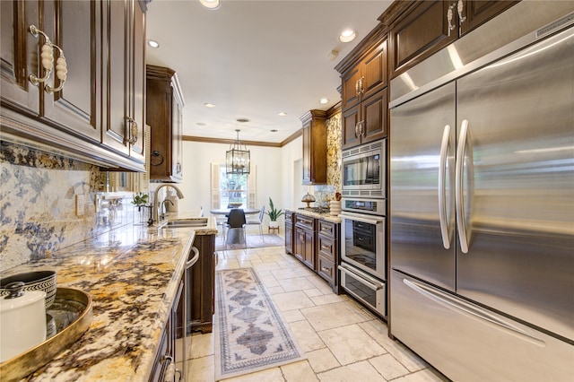 kitchen with light stone counters, ornamental molding, sink, pendant lighting, and built in appliances