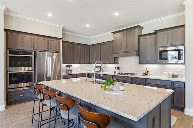 kitchen with a kitchen island with sink, stainless steel appliances, light stone countertops, ornamental molding, and light hardwood / wood-style floors