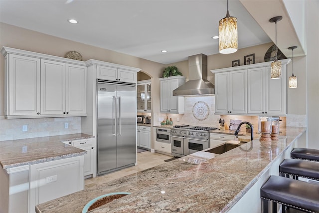 kitchen with high end appliances, kitchen peninsula, sink, wall chimney exhaust hood, and white cabinetry