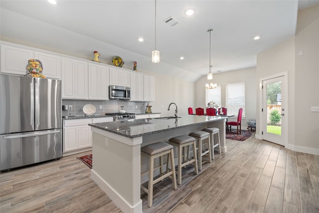 kitchen featuring appliances with stainless steel finishes, light wood-type flooring, a center island with sink, dark stone countertops, and white cabinetry