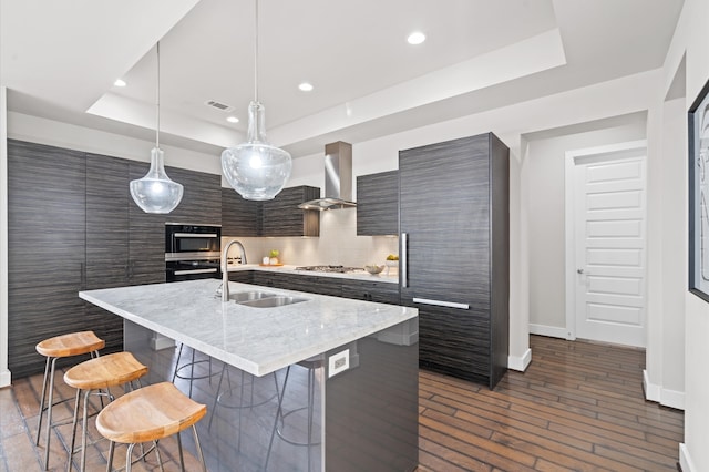 kitchen with a raised ceiling, wall chimney range hood, a center island with sink, hanging light fixtures, and a breakfast bar area