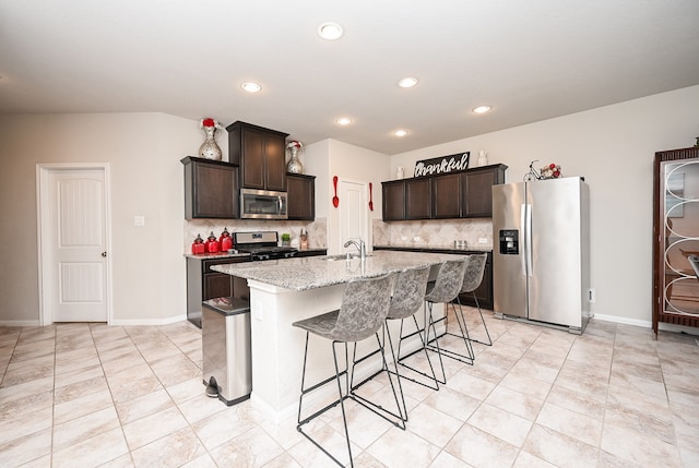 kitchen with light stone countertops, appliances with stainless steel finishes, backsplash, dark brown cabinets, and a kitchen island with sink