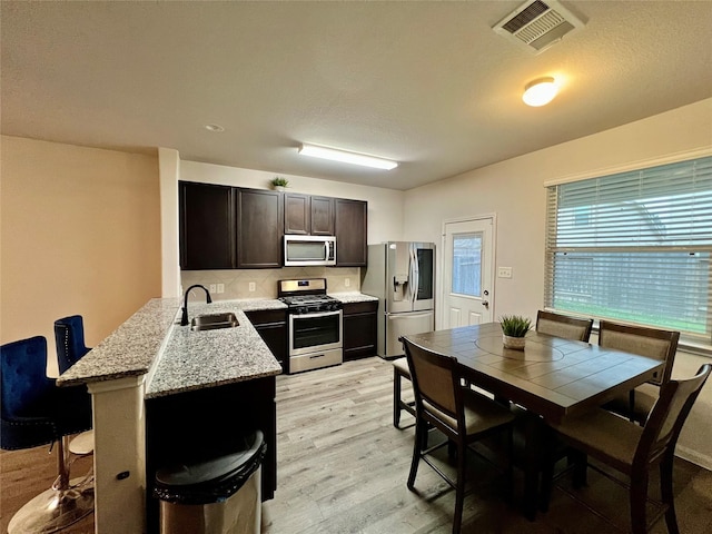 kitchen with appliances with stainless steel finishes, light wood-type flooring, light stone counters, dark brown cabinets, and sink