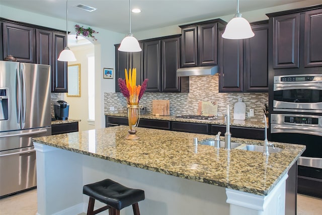 kitchen featuring light stone counters, stainless steel appliances, sink, hanging light fixtures, and an island with sink