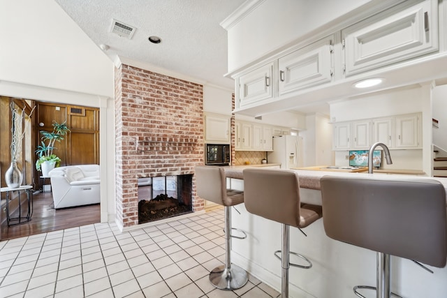 kitchen featuring a brick fireplace, white fridge with ice dispenser, a textured ceiling, a kitchen bar, and white cabinetry