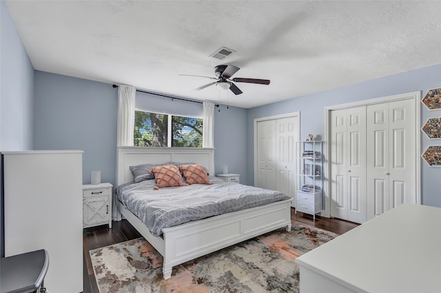 bedroom with a textured ceiling, two closets, ceiling fan, and dark wood-type flooring