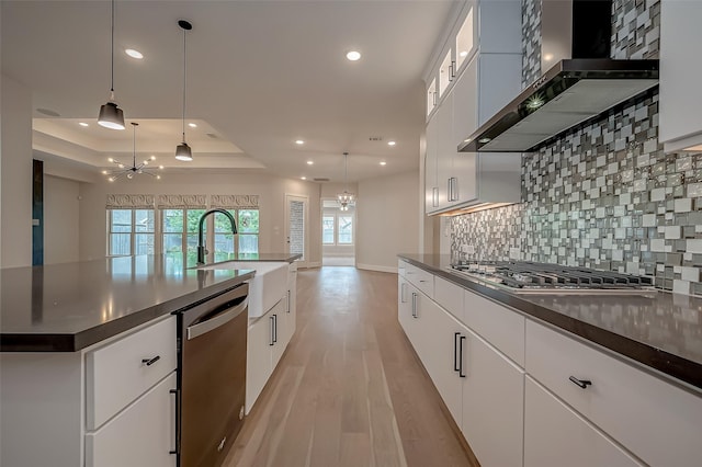 kitchen with appliances with stainless steel finishes, a tray ceiling, sink, wall chimney range hood, and an inviting chandelier