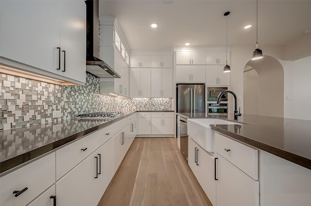 kitchen with stainless steel appliances, wall chimney range hood, white cabinets, light hardwood / wood-style floors, and hanging light fixtures