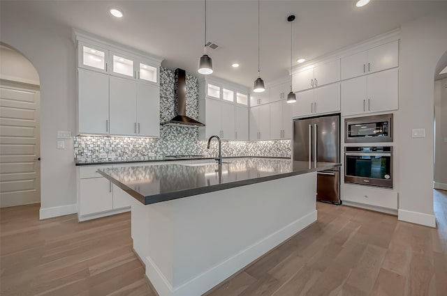 kitchen with white cabinetry, stainless steel appliances, wall chimney range hood, pendant lighting, and a center island with sink