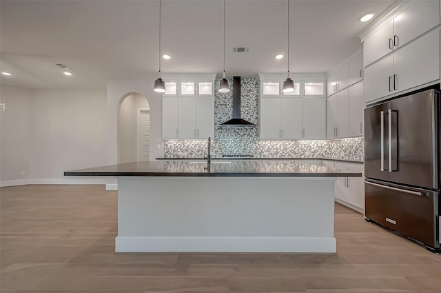 kitchen featuring high end refrigerator, a center island with sink, hanging light fixtures, wall chimney exhaust hood, and white cabinetry