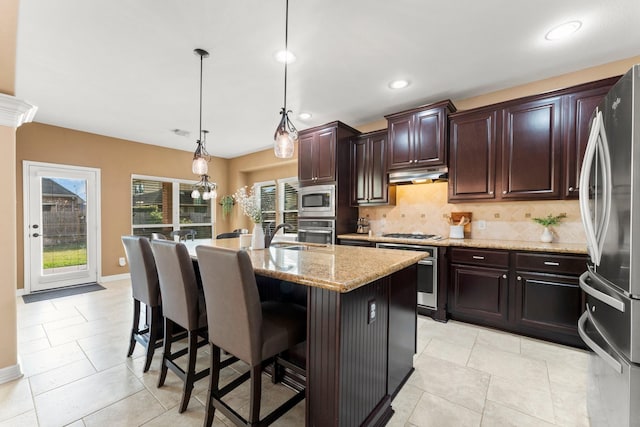 kitchen featuring a center island with sink, under cabinet range hood, light stone counters, tasteful backsplash, and appliances with stainless steel finishes