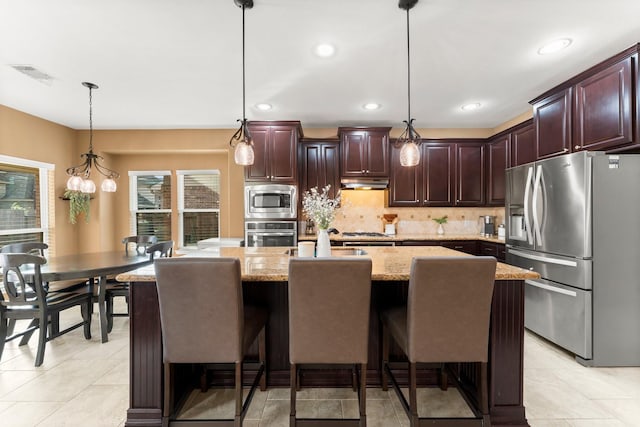 kitchen with appliances with stainless steel finishes, light stone counters, a kitchen island with sink, decorative light fixtures, and a breakfast bar area