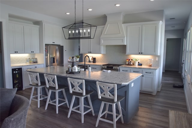 kitchen with white cabinets, custom range hood, a kitchen island with sink, and appliances with stainless steel finishes