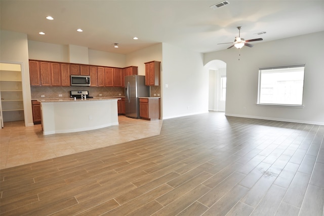 kitchen featuring ceiling fan, a kitchen island with sink, stainless steel appliances, light hardwood / wood-style flooring, and decorative backsplash