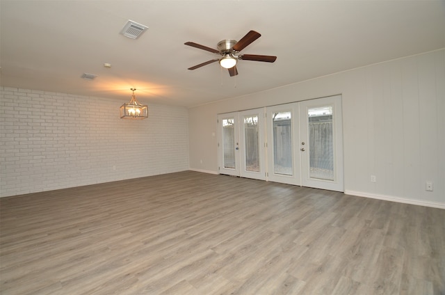 empty room featuring ceiling fan with notable chandelier, light hardwood / wood-style floors, brick wall, and french doors