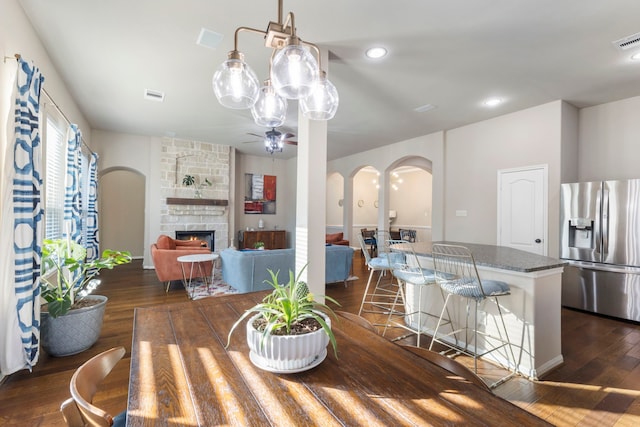 dining area featuring ceiling fan with notable chandelier, dark wood-type flooring, and a stone fireplace