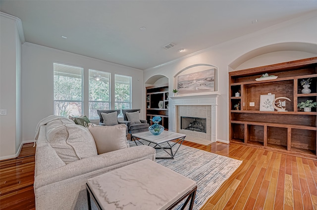 living room with built in shelves, a tiled fireplace, ornamental molding, and light hardwood / wood-style flooring